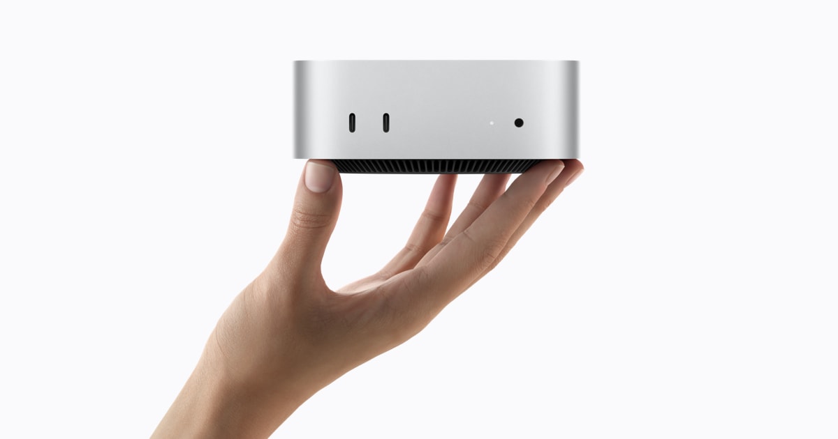 A compact silver Mac mini balanced on an outstretched hand against a white background, viewed from the front with two USB-C ports, a small white status light, a headphone jack, and black ventilation u
