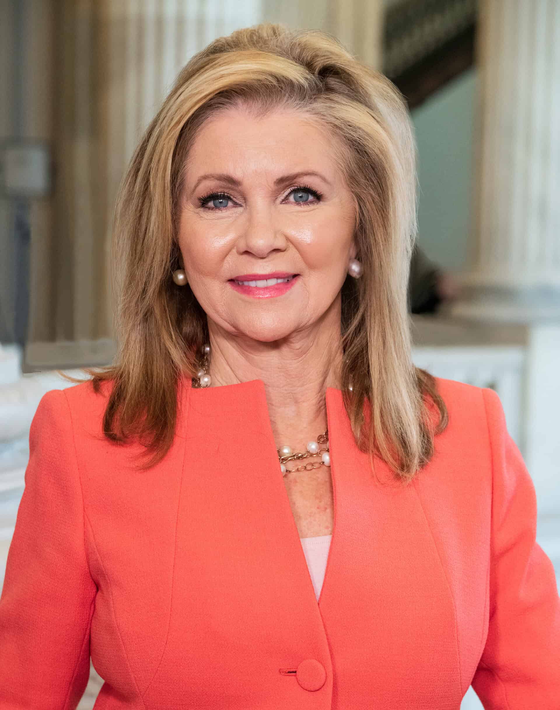 A real photographic portrait of U.S. Senator Marsha Blackburn in a coral blazer inside the U.S. Capitol.