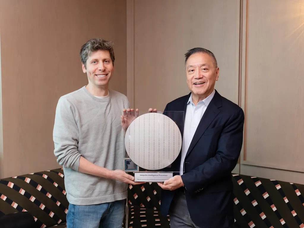 A posed indoor photograph of Sam Altman, at left in a gray sweater, and another man in a dark suit, apparently Richard, jointly holding a large circular semiconductor wafer mounted on a display labele