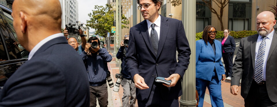 A tall man in a dark suit and glasses walks on a city sidewalk holding a phone or small device, surrounded by security personnel and photographers with large cameras. The image does not clearly establ